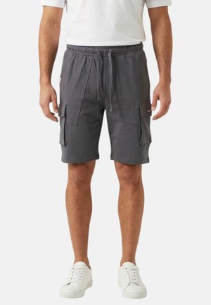Homme portant un short cargo gris avec taille à cordon et baskets blanches, debout devant un fond uni.