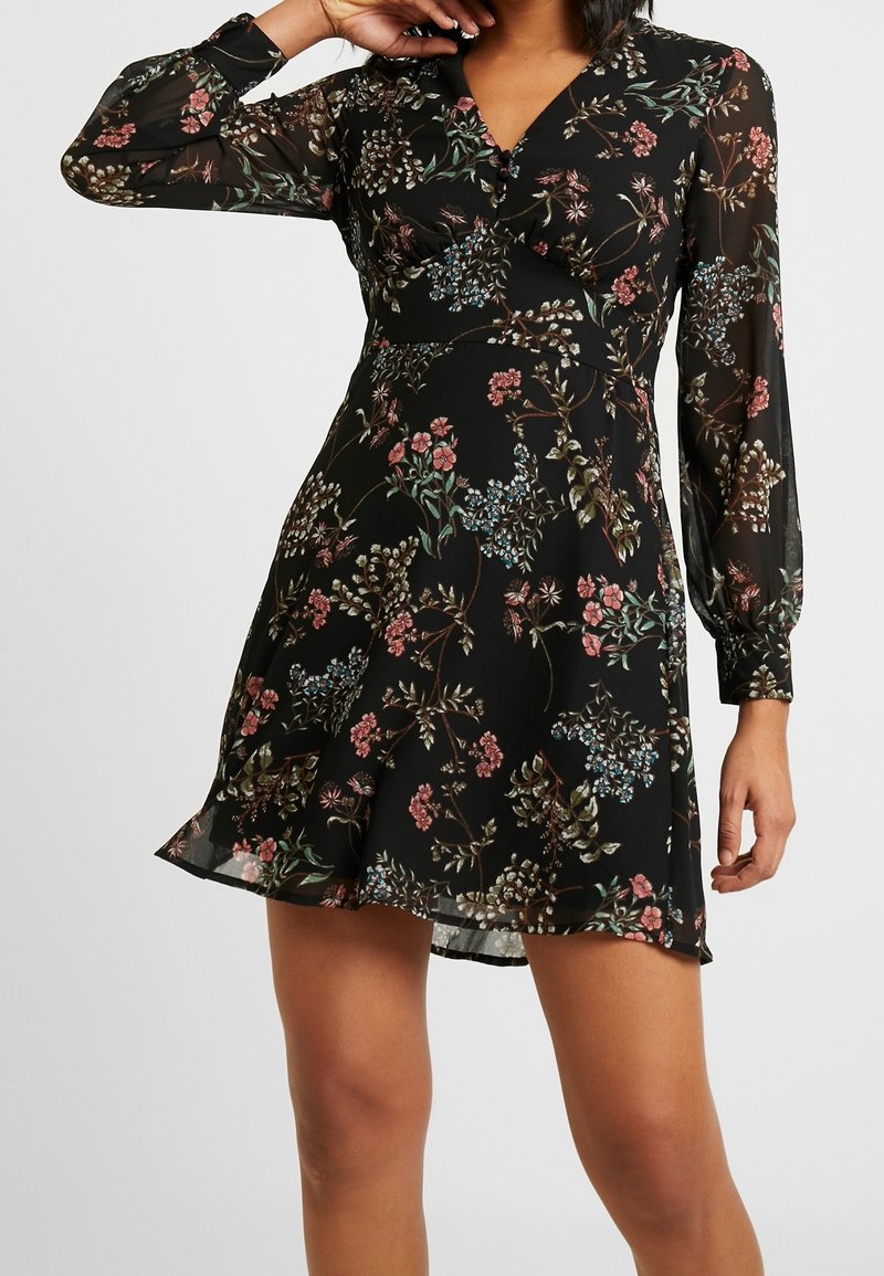 Femme portant une robe portefeuille noire à motif floral, avec de longues manches transparentes et une jupe évasée, sur un fond uni.