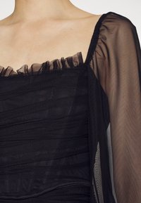 Robe noire transparente avec corsage froncé, encolure à volants et longues manches transparentes sur une épaule et un haut de poitrine à teint clair.