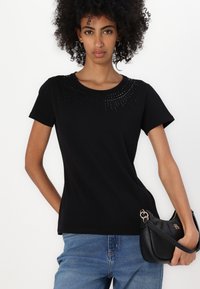 T-shirt nera a maniche corte con scollatura arrotondata, caratterizzata da un motivo decorativo di dettagli metallici al collo. Abbinata a jeans blu.