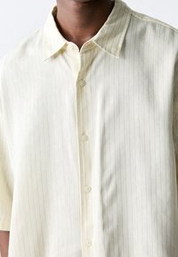 Koszula z krótkim rękawem w jasnym odcieniu żółtego, zapinana na guziki, z delikatnymi pionowymi paskami, klasycznym kołnierzykiem i gładką fakturą. Posiada dopasowane guziki.