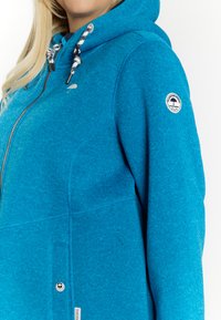 Felpa blu con zip realizzata in tessuto testurizzato, cappuccio con cordini decorati e patch del logo sulla spalla sinistra.