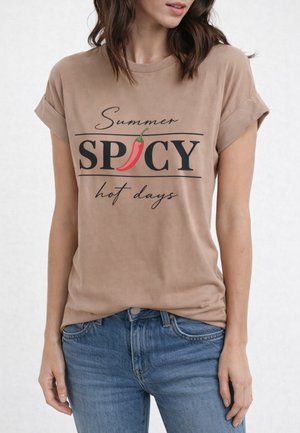 Donna con t-shirt color sabbia a maniche arrotolate con testo nero "Summer SPICY hot days" e una grafica di peperoncino rosso al posto della "I".