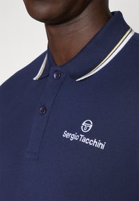 Námornícka polo tričko s rebrovaným golierom s bielymi a žltými pruhmi. Vyšívané logo v bielej farbe na hrudi, s fialovými gombíkmi.
