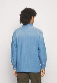 Chemise en denim bleu clair avec manches longues, poignets à boutons et empiècement dans le dos. Présente une texture subtile et une coupe décontractée.