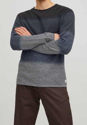 Mężczyzna noszący sweter z długim rękawem w kolorach granatowym i szarym oraz brązowe spodnie, z założonymi skrzyżowanymi ramionami, na jednolitym tle.