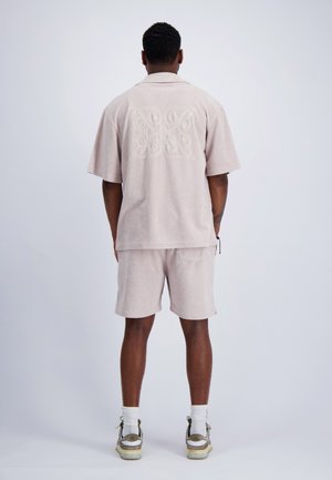 Man staat met zijn rug naar de camera, draagt een lichtbeige bijpassend kort mouwen overhemd en korte broek met witte sokken en sneakers tegen een effen achtergrond.