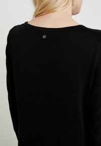 Černý pletený svetr s kulatým výstřihem a malou černou štítkovou nášivkou na zadní straně. Textura vypadá jemně a mírně žebrovaně.