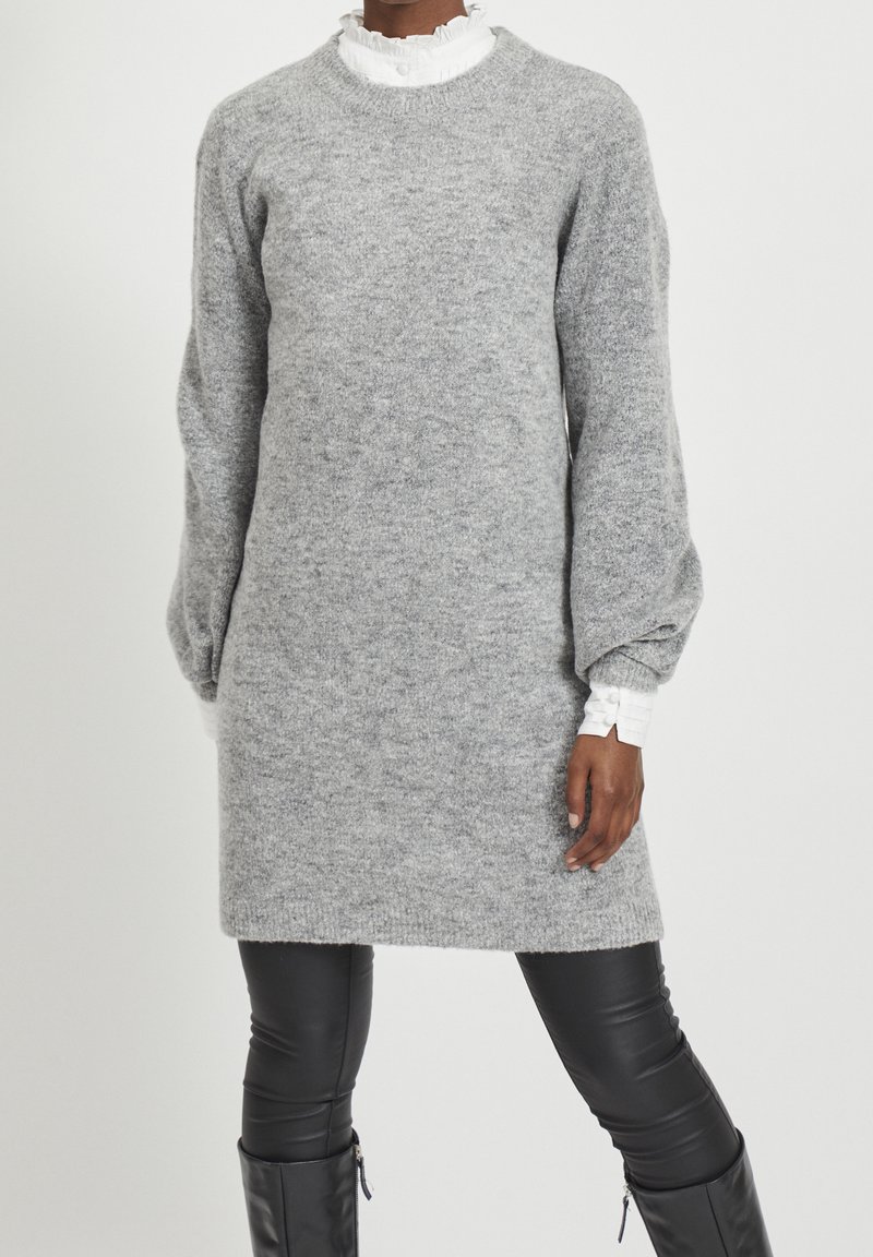 Abito maglione grigio realizzato in morbido materiale knit, con scollo tondo e maniche lunghe a sbuffo. Indossato sopra leggings in pelle nera.