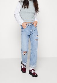 Jeans in denim chiaro con effetto vissuto e strappi, abbinati a una maglietta grigia a costine senza maniche e scarpe in pelle verniciata bordeaux.