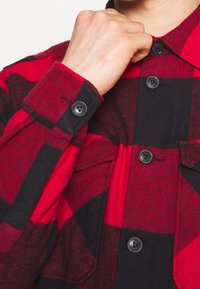 Camicia di flanella a scacchi rossi e neri con chiusura a bottoni, maniche lunghe e un tessuto morbido e texture con tasche sul petto.