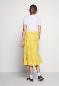 Kvinne med hår i en knute iført en hvid t-shirt, gul blomstrete midi-nederdel, hvide sokker og marineblå sneakers, stående foran en ensfarvet baggrund.