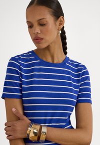 Žena s pletenými vlasmi, ktorá má na sebe modro-bielo pruhované tričko, zlaté kruhové náušnice a niekoľko zlatých a modrých náramkov na ruke.