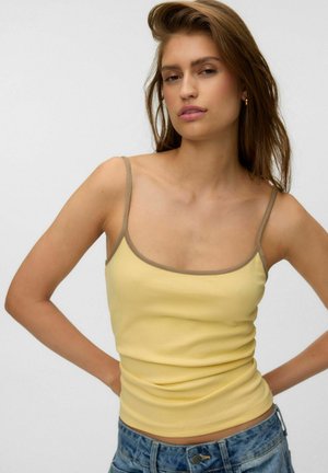 Jonge vrouw met lang bruin haar die een geel tanktopje met bruine bandjes en een blauwe spijkerbroek draagt, staat met handen in de zij tegen een effen achtergrond.