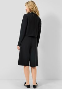 Sieviete ar viļņainiem gaišiem matiem stāv, ar muguru vērsta uz skatītāju, tērpta melnā īsajā jakā, platu garu šortu līdz ceļiem un melnām plakanām kurpēm.