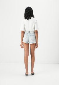 Haut blanc à manches courtes associé à un short en denim délavé bleu clair. Le short présente des ourlets effilochés et des poches arrière. Modèle portant des sandales à talons grises.