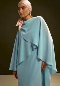 Vestido azul claro con diseño fluido tipo capa, con textura ondulada. Combinado con un pañuelo para la cabeza rosa pálido y pendientes plateados.