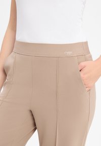 Beige Hose mit elastischem Bund, ausgestattet mit zwei Seitentaschen und einem glatten, leichten Stoff. Markenlogo über dem Bund gestickt.