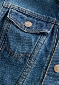 Détail d'une veste en denim avec un tissu bleu clair, des coutures orange contrastantes, une poche à rabat et un bouton logo en argent marqué "AUTHENTIC 1982."