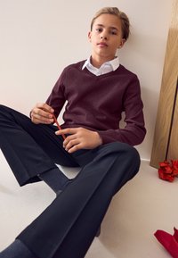 Bordó V-nyakú pulóver fehér galléros ing fölött, fekete nadrággal párosítva. Ülő testtartás, narancssárga ceruzát tart, szürke zoknik láthatók.