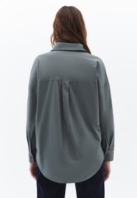 Camicia grigia chiaro a maniche lunghe con colletto classico e orlo arrotondato, caratterizzata da una piega centrale sul retro e polsi con bottoni. Tessuto liscio.
