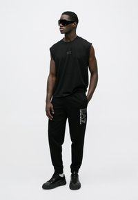 Fekete ujjatlan póló EA7 logóval, fekete logóval díszített melegítőnadrággal párosítva. Az összeállítást fekete sportcipő teszi teljessé.