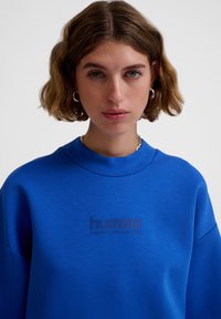 Blaues Sweatshirt mit rundem Ausschnitt, oversized Passform und weichem Material. Mit geprägtem "hummel PERFORMANCE"-Logo auf der Brust.