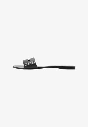 Black slide sandal with a flat sole, wide strap featuring a geometric cutout and metallic accents, crafted from smooth leather-like material.