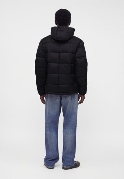 Homme portant une doudoune noire à capuche et un jean bleu large, debout de dos sur un fond clair et uni.