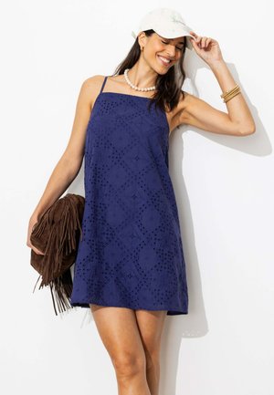 Femme en robe bleue sans manches avec motif œillet, casquette blanche, collier de perles, bracelets dorés, tenant un sac frangé marron, souriant les yeux fermés.