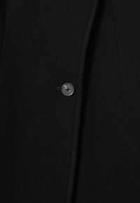 Schwarzer Wollblazer mit einem einzelnen grauen Knopf und einer glatten Textur, dazu dezente Nähte und ein taillierter Schnitt.