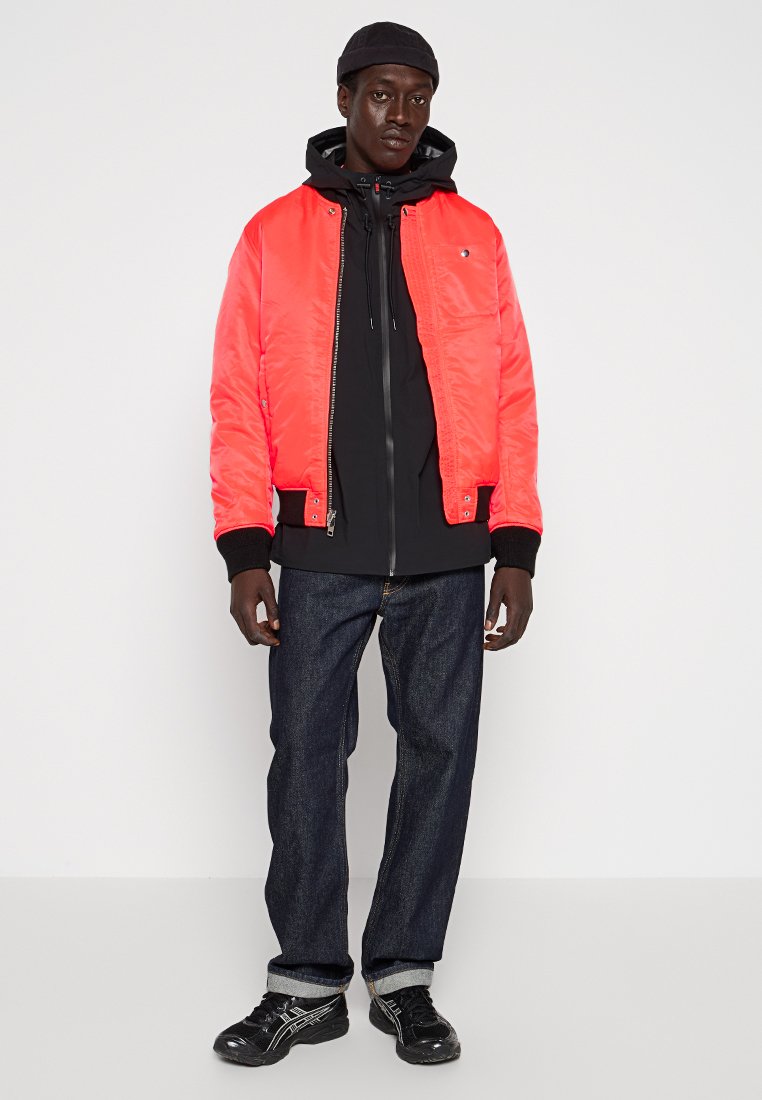 Homme portant une veste bomber rouge vif sur un hoodie noir, un jean sombre, des baskets noires et une casquette noire, se tenant devant un fond blanc.