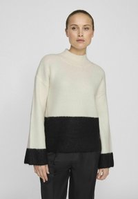 Ljust beige ribbat tröja med en svart nedre del och ärmar. Hög halsringning, avslappnad passform och mjuk textur. Modellen har matchat med mörka byxor.
