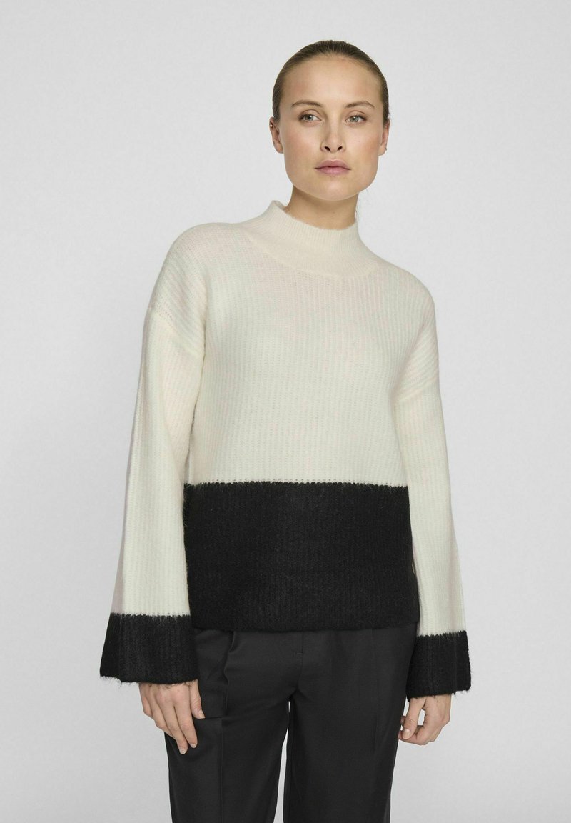 Ljust beige ribbat tröja med en svart nedre del och ärmar. Hög halsringning, avslappnad passform och mjuk textur. Modellen har matchat med mörka byxor.