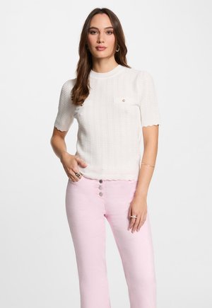 Maglione bianco lavorato a maglia con maniche corte e orlo a scallop, dotato di due piccole tasche sul petto e un delicato motivo a texture; abbinato a pantaloni rosa.