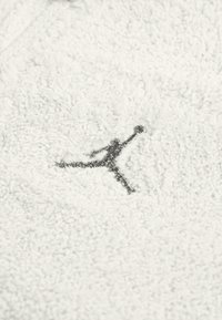 Biała, fakturowana odzież z polaru z szarym haftowanym logo Jumpman. Materiał wygląda na miękki i przytulny, z delikatnym wzorem na powierzchni.