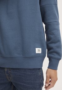 Blauwe sweatshirt met ribgebreide manchetten en onderrand, gemaakt van een katoenmix. Bevat een klein wit label met zwarte tekst onderaan.