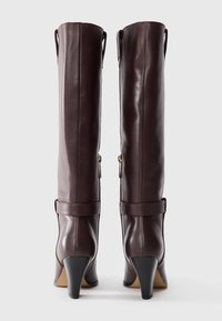 Stivali in pelle bordeaux alti fino al ginocchio, con una forma aderente, punta appuntita e tacco a stiletto nero. Presentano un cinturino e una chiusura con zip.
