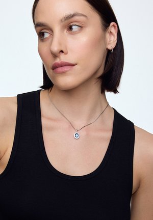 Srebrny naszyjnik z okrągłym zawieszeniem w niebieskim kolorze i białymi akcentami, zawieszony na cienkim metalowym łańcuszku, noszony z czarnym topem na ramiączkach.