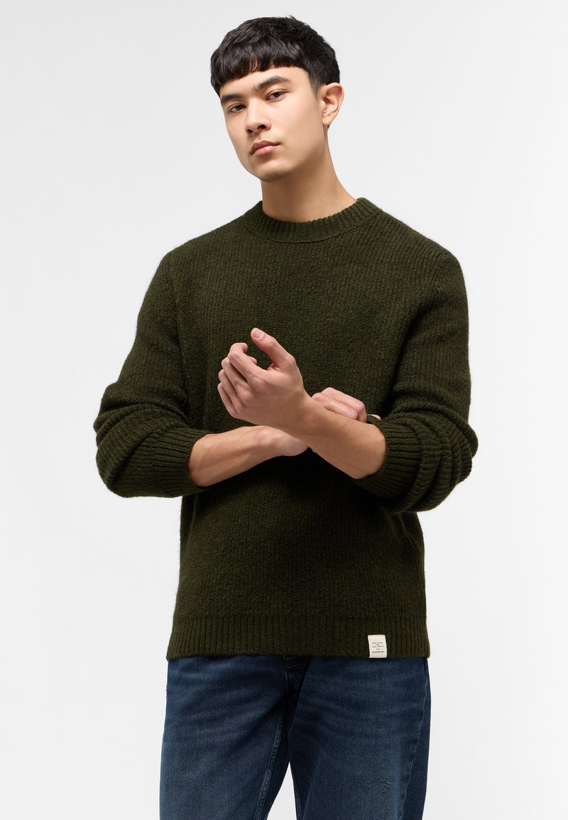Suéter de punto verde oscuro con un diseño de canalé texturizado, cuello redondo y puños enrollados. Combinado con jeans azules, que tienen una etiqueta en la parte inferior.