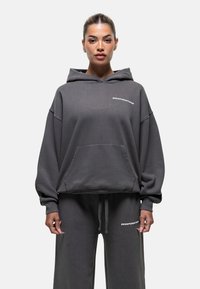 Haut à capuche surdimensionné gris en tissu doux, avec une poche avant et un logo blanc sur la poitrine, associé à un pantalon de jogging assorti.