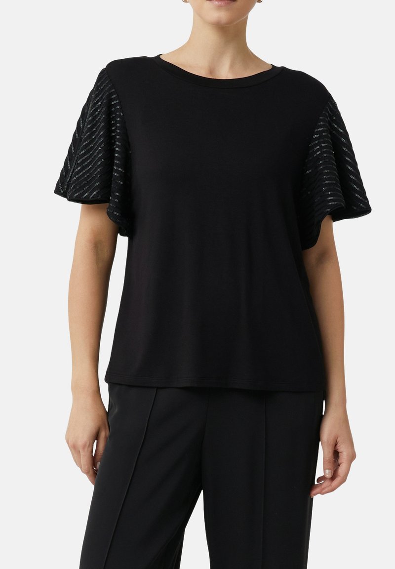 Frau trägt ein schwarzes, weit geschnittenes Oberteil mit strukturierten, gestreiften kurzen Schmetterlingsärmeln und schwarze, maßgeschneiderte Hosen, steht vor einem weißen Hintergrund.