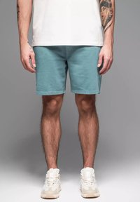 Shorts turquoise en tissu doux, avec une coupe droite et un ourlet soigné. Portés avec des baskets blanches. Arrière-plan gris.