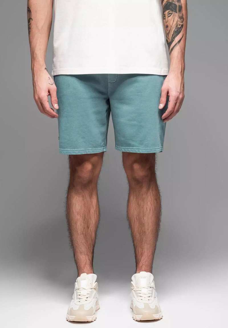 Shorts turquoise en tissu doux, avec une coupe droite et un ourlet soigné. Portés avec des baskets blanches. Arrière-plan gris.