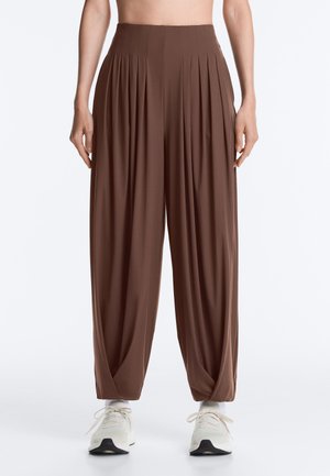 Pantalon marron plissé taille haute à jambes larges, porté avec des chaussettes blanches et des baskets blanches à lacets, debout sur un fond uni.