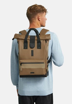 Brauner Rucksack mit Klappe und schwarzen Akzenten sowie einer vorderen Reißverschlusstasche. Hergestellt aus strapazierfähigem Material, mit verstellbaren Gurten und seitlichen Clips.