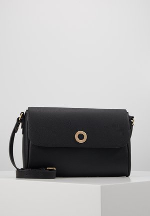 Sac bandoulière en cuir noir avec surface texturée, fermeture à rabat, fermoir circulaire doré et sangle réglable sur une surface blanche contre un fond gris clair.