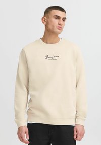 Giovane uomo vestito con una felpa beige con la scritta "Bonjour Côte d'Azur", abbinata a pantaloni neri, in piedi su uno sfondo chiaro e uniforme.