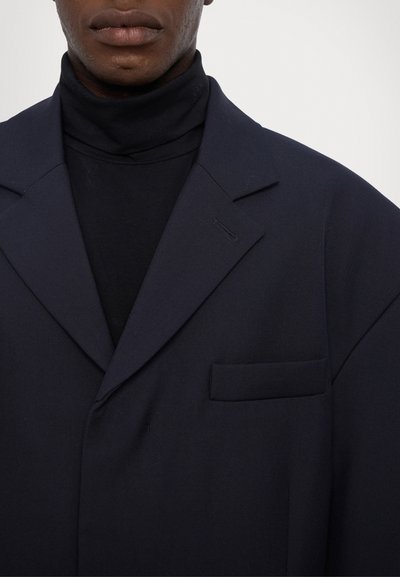 Marineblazer mit ausgeprägten Revers, einer einzelnen Brusttasche und einer glatten Stoffstruktur, über einem formschlüssigen schwarzen Rollkragenpullover getragen.