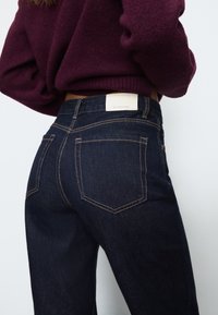 Temno modri jeans z oprijetim krojem, ki imajo kontrastne oranžne šive, dve zadnji žepki in belo oznako blagovne znamke na pasu.